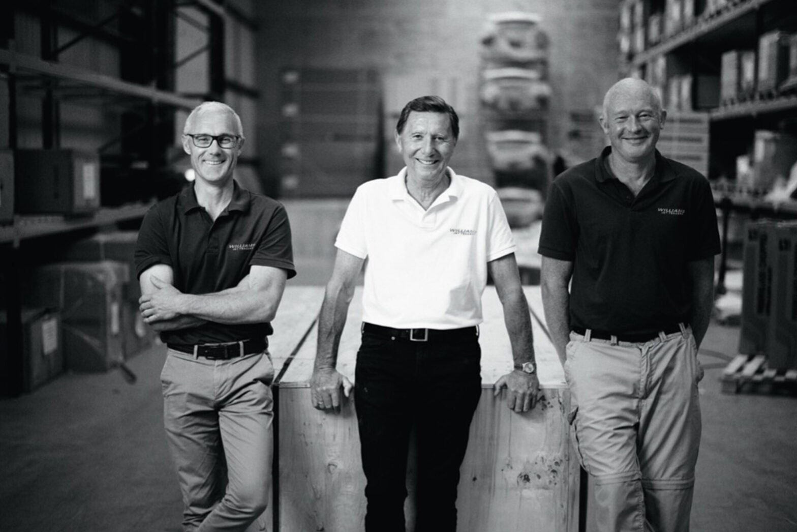 Three men in Williams Jet Tenders shirts are standing in a warehouse, smiling confidently